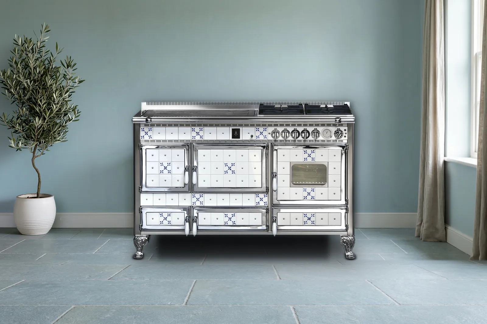 Cuisinière mixte Archambaud Origine 140 en faïence blanche à motifs bleus et chrome dans une cuisine de campagne, mur bleu-gris et dalles de pierre